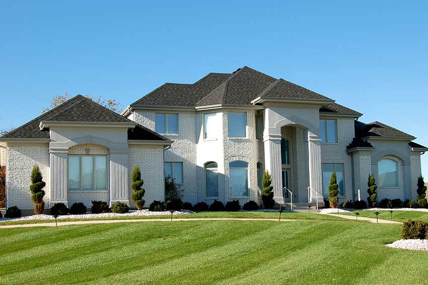 Luxurious two-story brick house with dark roof, elegant columns, and manicured lawn under a clear blue sky, conveying a sense of grandeur and serenity.
